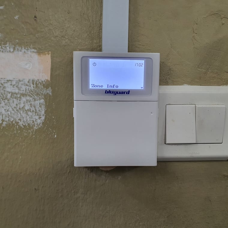 A Bluguard alarm keypad showing a Zone Info screen, mounted beside light switches.