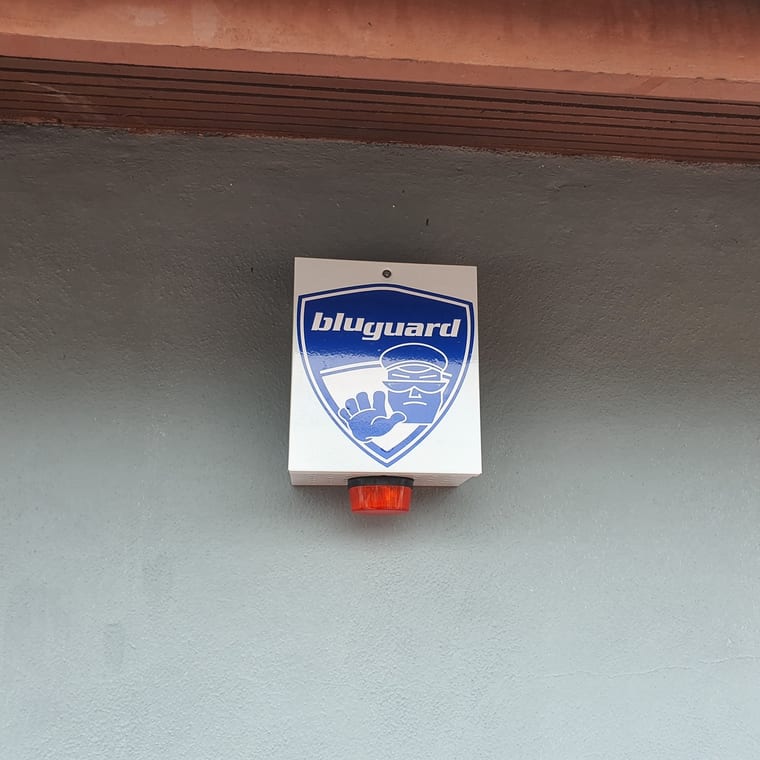 A Bluguard outdoor siren housing with a red strobe lens, fixed high on an exterior wall under a tiled eave.