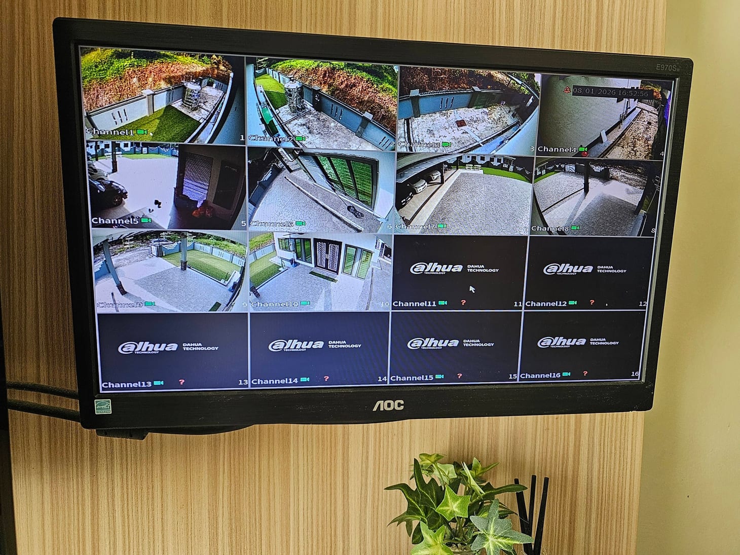 A wall-mounted monitor showing a sixteen-tile CCTV live view of a house's garden, driveway, gate and exterior walls.