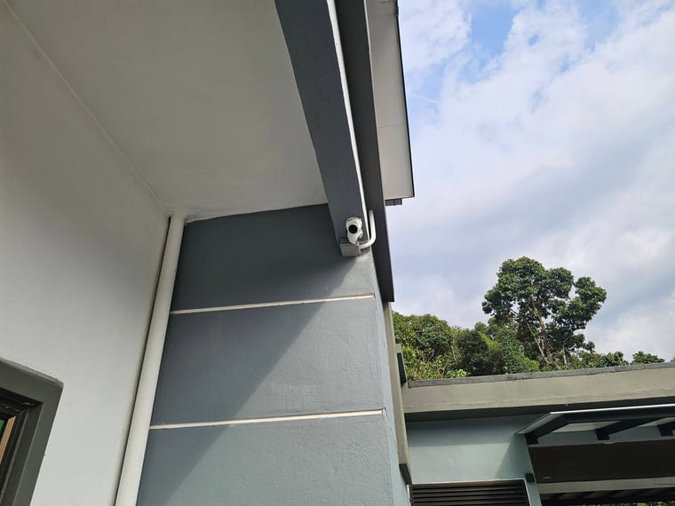 A camera fixed beneath a dark eave, seen from below against an overcast sky.