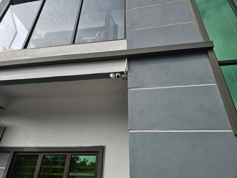 A camera on a dark beam against a grey-panelled house facade, with a window and trees behind.
