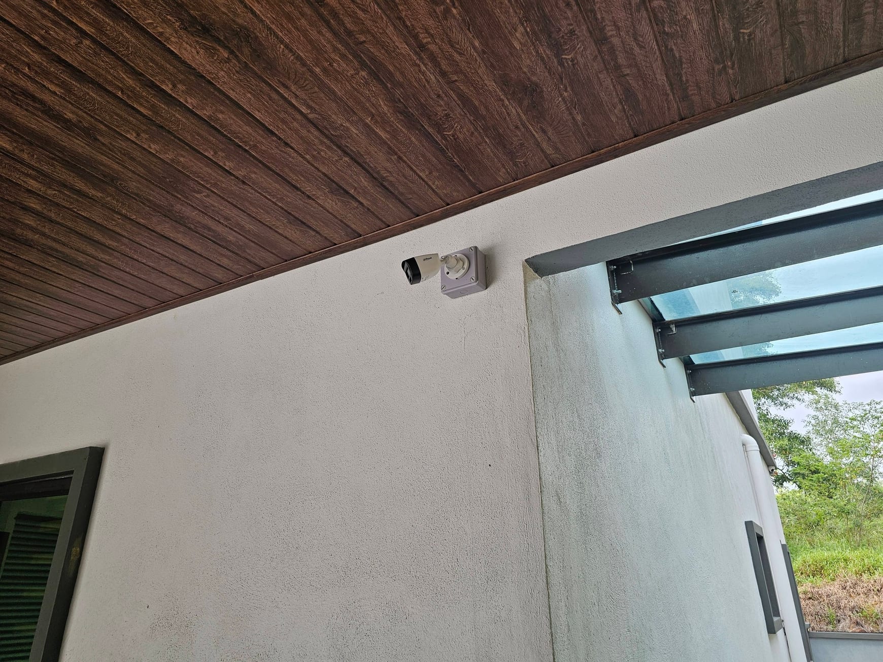 A bullet camera fixed to a dark timber-clad soffit above a glazed garden door, with foliage beyond.